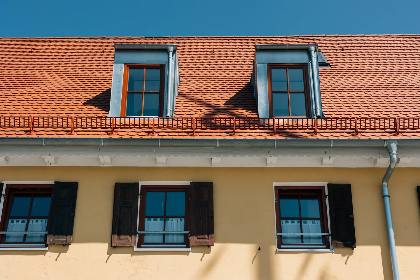 Zwei Dachgauben mit Fenstern über einer gelben Fassade, darunter Fenster mit dunklen Fensterläden.