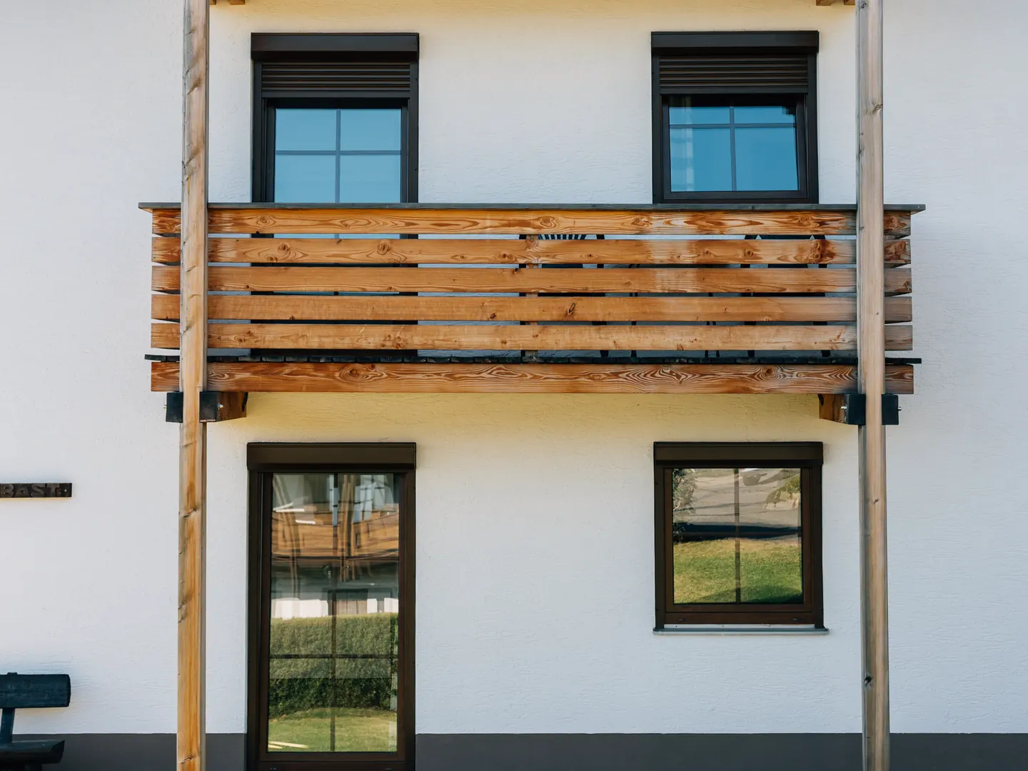 Weiße Fassade mit Holzbalkon, zwei Fenstern mit Rollladenkästen und einer Balkontür im Erdgeschoss.