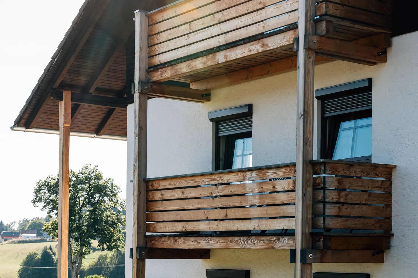 Dunkle Fenster mit außen liegenden Rollladenkästen an einer hellen Hauswand, davor Holzbalkone.