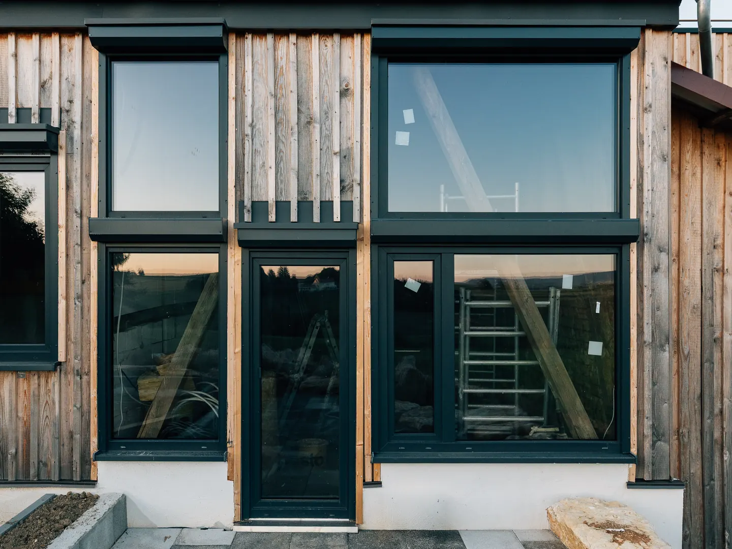 Dunkle Fenster und eine verglaste Tür in einer Holzfassade, auf den Scheiben noch Aufkleber.