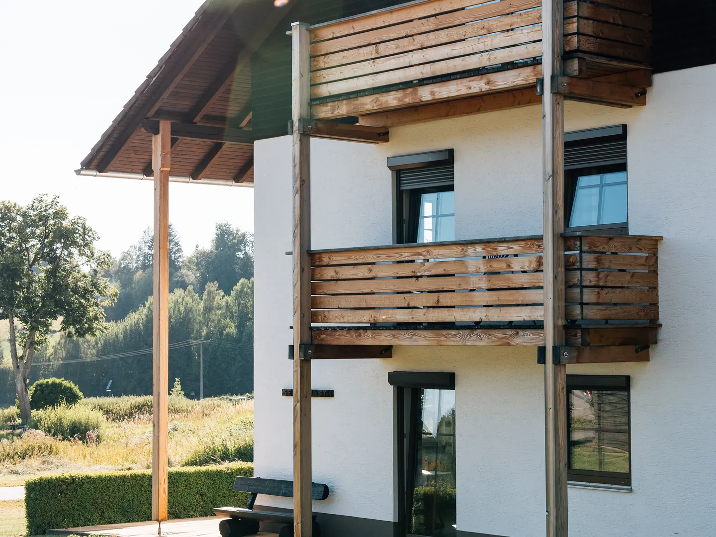 Weiße Hausfassade mit Holzbalkon, dunklen Fenstern mit Rollläden und einer Fenstertür im Erdgeschoss.