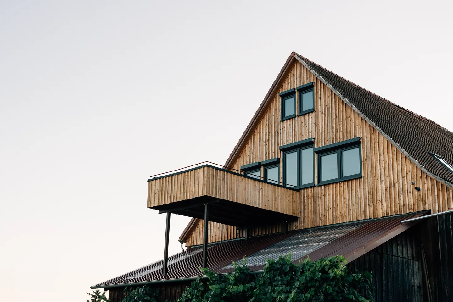 Giebel mit senkrechter Holzverschalung, dunklen Fenstern und einem Balkon vor hellem Himmel.
