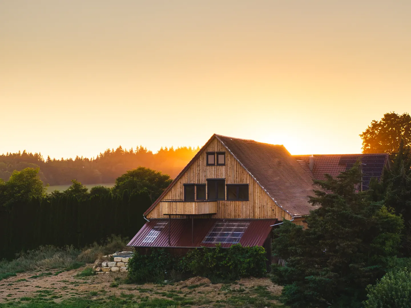Holzhaus mit Satteldach und Fenstern im Giebel im Abendlicht, dahinter Wald und tief stehende Sonne.
