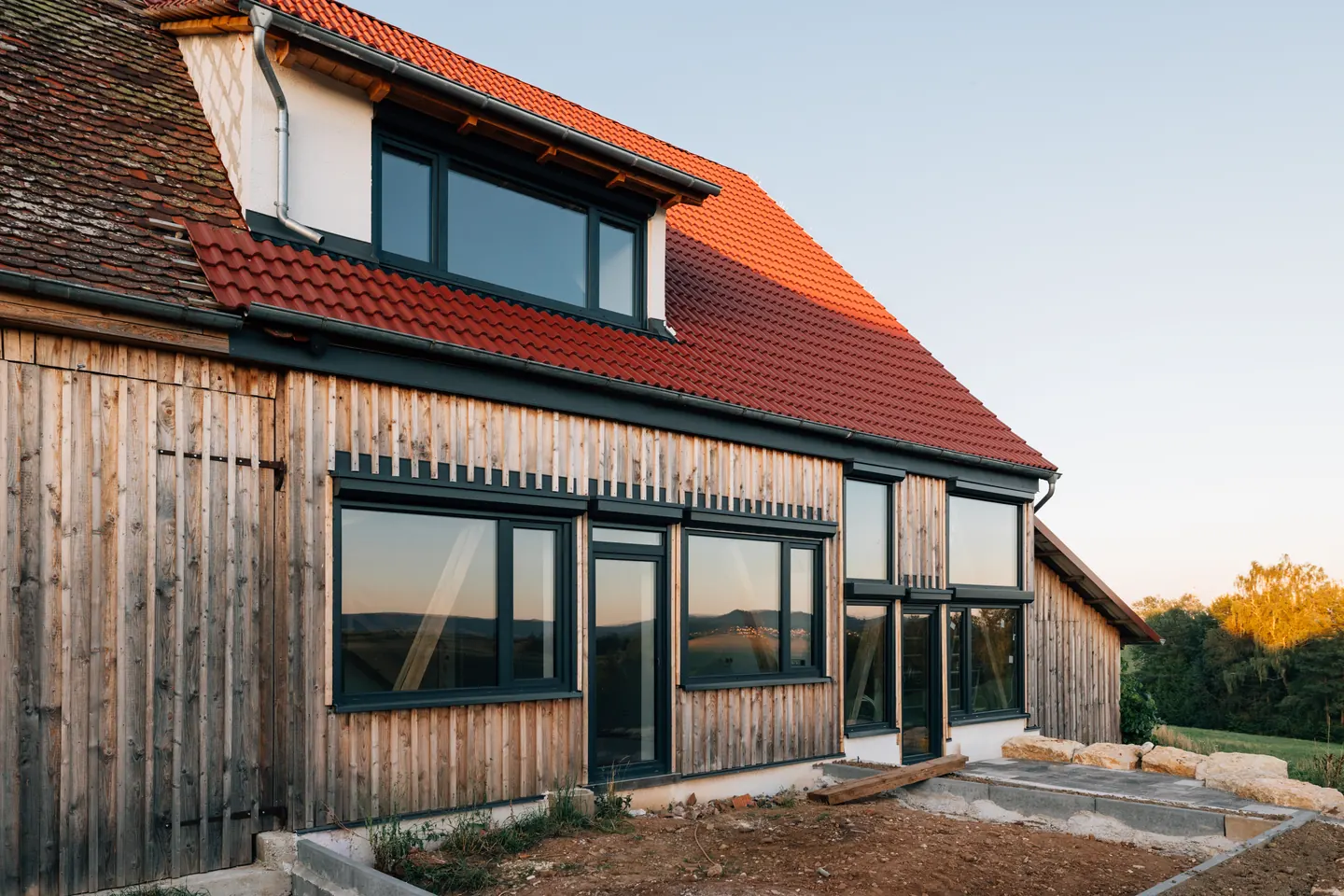 Holzhaus mit senkrechter Holzverschalung und großen dunklen Fenstern, darüber ein rotes Ziegeldach mit Gaube.