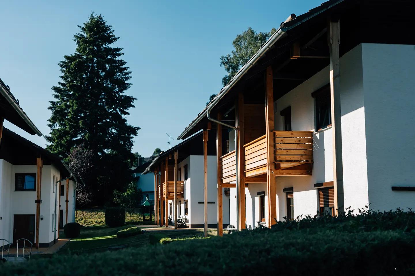 Wohnhäuser mit Holzbalkonen und Fenstern im Erdgeschoss, davor Garten und eine hohe Tanne.