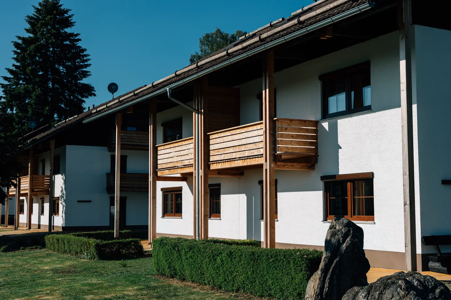 Weißes Wohnhaus mit Holzbalkonen und Fenstern mit Rahmen in Holzfarbe, davor eine Hecke.